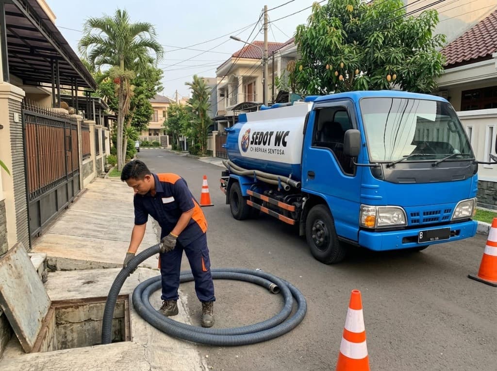 Teknisi Sedot WC Berkah memasang selang di saluran rumah, dengan truk tangki biru di belakang.